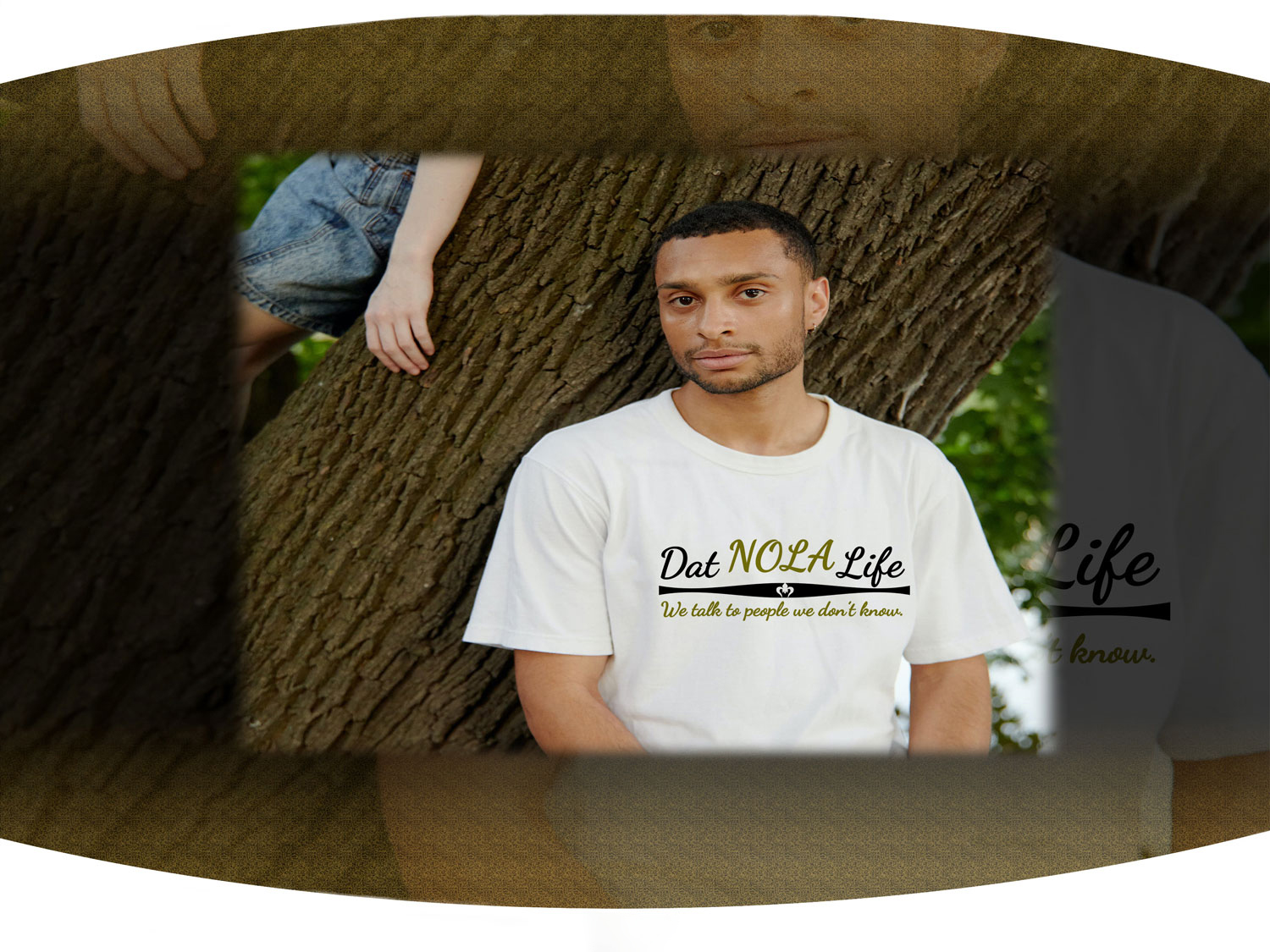 Man leaning on tree wearing white t-shirt with the black and gold Dat Nola Life text across the chest